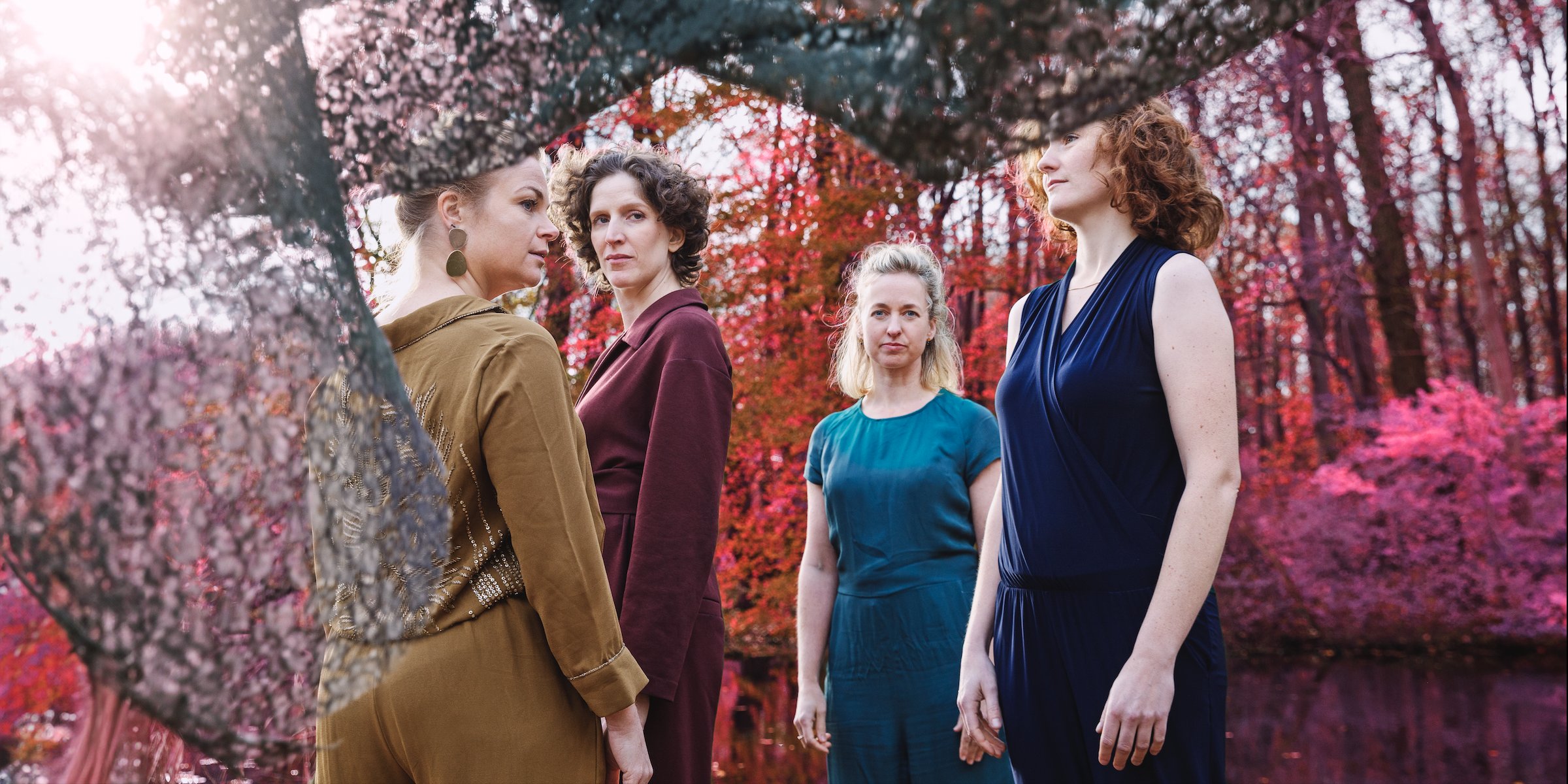 Four women (Ragazze Quartet) in color-blocked outfits in earthy tones. In the background are bright orange and pink trees and bushes.